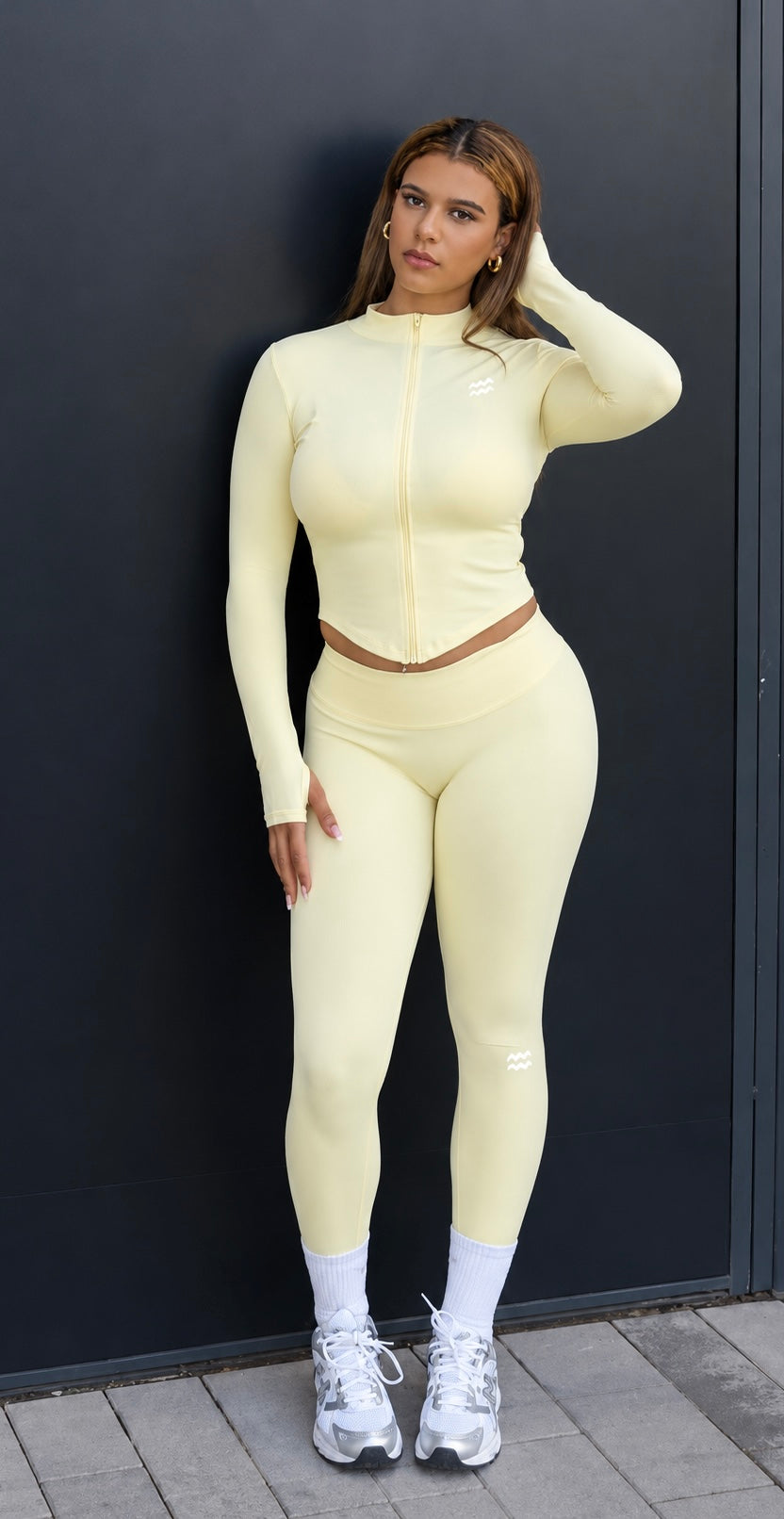 A woman modeling a pale yellow matching two-piece activewear set consisting of a long-sleeve zip-up top and high-waisted leggings, styled with white socks and white sneakers against a dark gray wall.